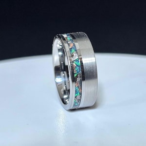 May include: A silver ring with a brushed metal finish. The ring features a central inlay of shimmering green and blue material, creating a striking contrast. The ring is set against a white surface.