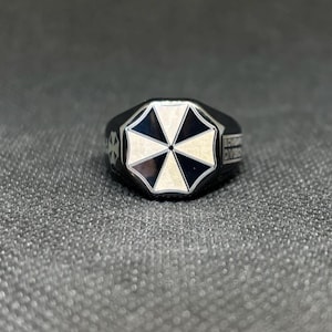 Puede incluir: Anillo negro con un emblema blanco y negro en forma de paraguas en la cara. El anillo tiene un dise&ntilde;o geom&eacute;trico y las palabras "SCORPORATION DIVISION" est&aacute;n grabadas en el lateral.