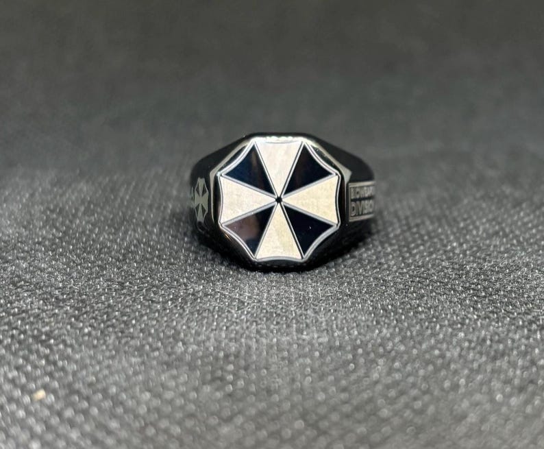 Puede incluir: Anillo negro con un emblema blanco y negro en forma de paraguas en la cara. El anillo tiene un dise&ntilde;o geom&eacute;trico y las palabras "SCORPORATION DIVISION" est&aacute;n grabadas en el lateral.