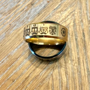 May include: Two rings, one gold-toned and one silver-toned, are displayed. The gold ring features etched images of Stormtrooper helmets and a symbol. The rings are set against a wooden surface.