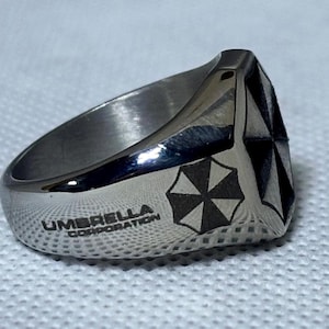 Puede incluir: Anillo plateado con el texto "UMBRELLA CORPORATION" y un logotipo de paraguas en blanco y negro. El anillo tiene un dise&ntilde;o geom&eacute;trico en la parte superior. Probablemente de acero inoxidable.