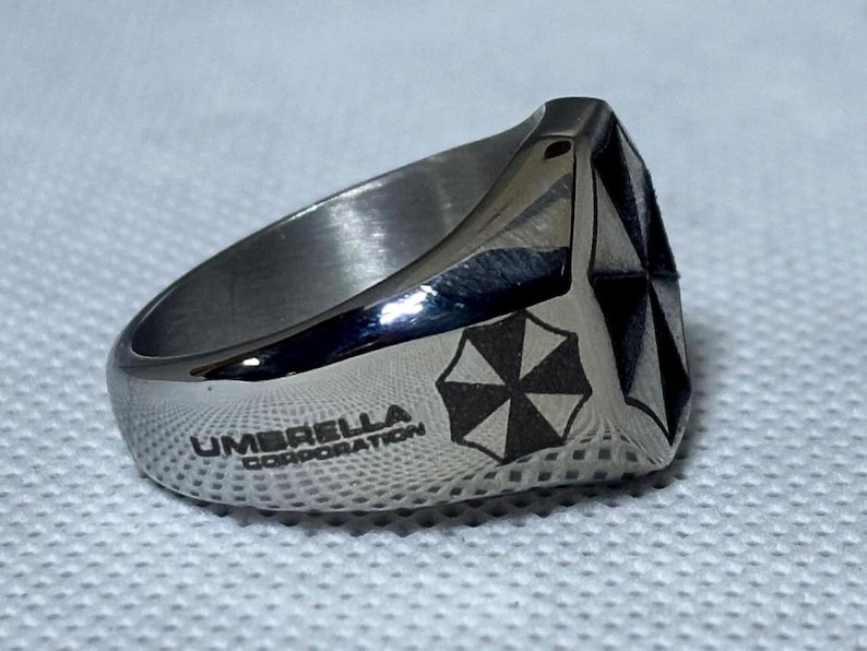 Puede incluir: Anillo plateado con el texto "UMBRELLA CORPORATION" y un logotipo de paraguas en blanco y negro. El anillo tiene un dise&ntilde;o geom&eacute;trico en la parte superior. Probablemente de acero inoxidable.