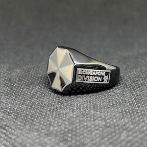 Puede incluir: Un anillo negro con un logotipo de paraguas plateado. El anillo tiene las palabras "BIOWEAPONS DIVISION" en texto blanco. El anillo est&aacute; sobre una superficie gris texturizada.