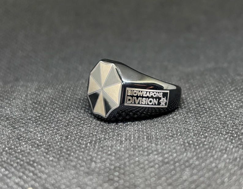 Puede incluir: Un anillo negro con un logotipo de paraguas plateado. El anillo tiene las palabras "BIOWEAPONS DIVISION" en texto blanco. El anillo est&aacute; sobre una superficie gris texturizada.