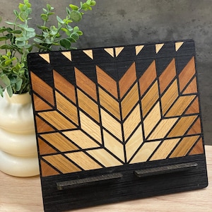 May include: A wooden phone stand with a geometric design in shades of brown and black. The stand has two slots for holding phones or tablets.