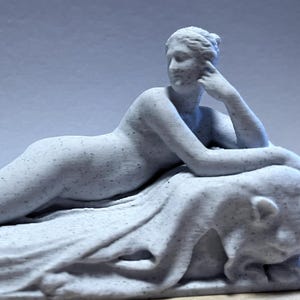 Puede incluir: Una escultura gris claro de una figura femenina reclinada, posiblemente una diosa clásica, descansando sobre una tela drapeada. La figura está en una pose relajada, con un brazo sosteniendo la cabeza. La escultura es detallada y parece ser de piedra.
