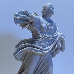 May include: A light gray statue of a draped figure with flowing fabric. The sculpture is standing on a rectangular base, with the figure's arms outstretched, and the fabric appears to be blowing in the wind. The statue has a classical aesthetic.