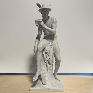 May include: A gray statue of a seated figure playing a pan flute. The figure wears a winged hat and is draped with fabric. The statue is on a pedestal base and is a classical art piece.