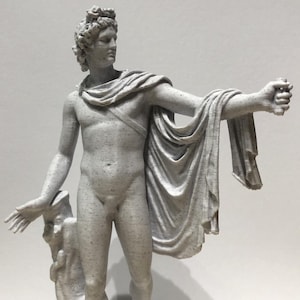 Puede incluir: Estatua detallada de una figura masculina, probablemente un dios clásico, con una tela drapeada sobre los hombros y una mano levantada. La estatua es de color gris claro con una textura moteada y se alza sobre una pequeña base.