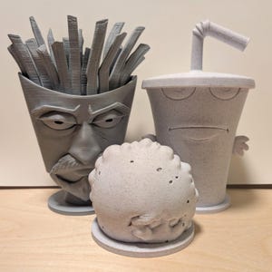 May include: Three 3D-printed gray figures representing a container of french fries with a stern face, a cup with a straw and a sleepy expression, and a round textured meatball-like character, all standing on circular bases.