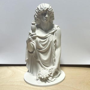 May include: A matte white 3D printed bust of a figure with curly hair, wearing a draped garment. The figure holds a small, pointed spindle-like object in the right hand and a circular, sun-shaped emblem in the left hand, set on a round base.