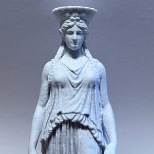 May include: A light gray speckled statue of a figure wearing a draped garment and a basket-like headpiece. The figure has braided hair and a calm facial expression. The material has a textured, stone-like appearance with fine dark flecks.