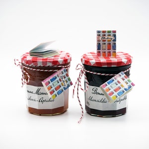 May include: Two glass jars of jam with red and white checkered lids. The jar on the left is filled with apricot jam and has a label that reads "Bonne Maman Abrikosi-Aprikos". The jar on the right is filled with blackberry jam and has a label that reads "Bonne Mansikka", with a small book on top of each jar.