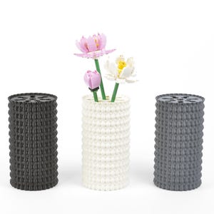 May include: Three cylindrical vases with a textured, dotted pattern. One is black, one is white, and the third is grey. The white vase holds artificial flowers in pink and white. The vases are decorative.