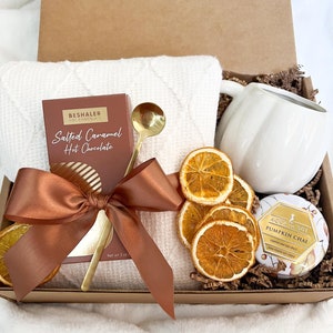 May include: A gift box filled with a white mug, a box of salted caramel hot chocolate, a gold spoon, a white waffle-weave towel, dried orange slices, a pumpkin spice candle, and a brown satin bow. The box is brown and filled with brown crinkle paper.