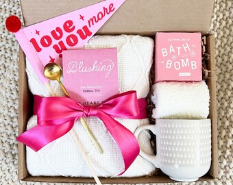 May include: A gift box filled with self-care items, including a white knitted blanket, a pink bath bomb, a mug, a pink tea bag, a gold spoon, and a pink pennant that says "love you more".