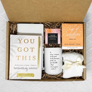 Op de afbeelding: Een cadeaudoos gevuld met artikelen voor zelfzorg en inspiratie. De doos bevat een witte mok met een citaat over kracht en aanmoediging, een boek getiteld "You Got This" met 90 devoties om hardwerkende vrouwen uit te rusten en te empoweren, een klein doosje thee met het label "Cup of Sunshine" met gouden kurkuma en gember, een witte handdoek met een gouden lint en een klein doosje badzout met het label "You're Wonderful".