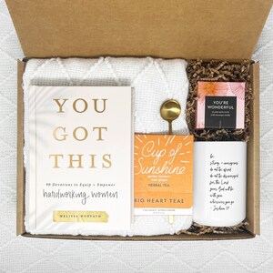 Op de afbeelding: Een cadeaudoos gevuld met artikelen voor zelfzorg en aanmoediging. De doos bevat een wit deken, een boek getiteld "You Got This" met de ondertitel "90 Devotions to Equip + Empower Hardworking Women", een doos kruidenthee met het label "Cup of Sunshine", een klein doosje met inspirerende kaarten en een witte mok met een citaat uit Jozua 1:9.