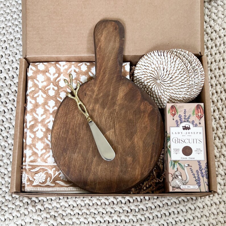 A brown wooden round cutting board with a handle, a gold metal butter knife, a brown and white patterned fabric napkin, a set of white woven coasters, and a box of Lady Joseph Cocoa Cacao biscuits.