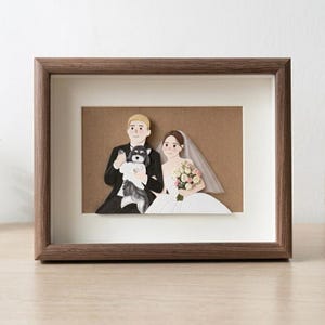 May include: A framed paper art piece featuring a 3D illustration of a wedding couple and a small dog. The person on the left wears a black suit, the person on the right wears a white dress and veil, and they hold a bouquet of pink and white flowers.