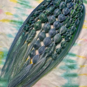 May include: A set of synthetic hair extensions featuring a mix of blue, green, and teal strands. The hair is styled into multiple bubble braids and fishtail braids, incorporating shimmering metallic fibers throughout the textured design.