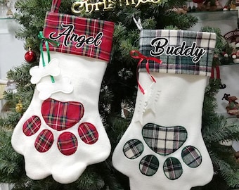 May include: Two white Christmas stockings shaped like a paw print, with red and green plaid accents. One stocking says "Angel" and the other says "Buddy". The stockings are hanging on a Christmas tree.