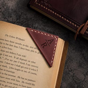 May include: A triangular dark brown leather corner bookmark featuring light-colored stitching along the edges and the embossed letters A and R separated by a horizontal line. The bookmark is placed on the corner of an open book page.
