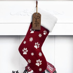 May include: A red Christmas stocking with white paw prints and snowflakes. The stocking has a white cuff and a brown tag that says "Justin". The stocking is hanging on a white mantel.