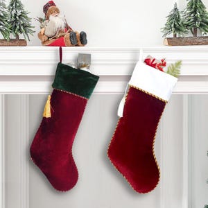May include: Two Christmas stockings in a festive display. One stocking is burgundy with a green velvet cuff and gold tassel. The other is burgundy with a white cuff and gold trim. Both are hanging from a white mantel.