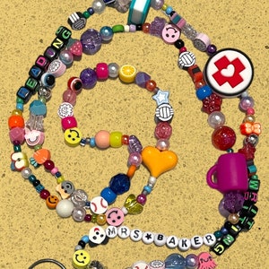 May include: A vibrant beaded lanyard with diverse charms, such as smiley faces, baseballs, and a miniature book. The lanyard spells out "READING" and "MRS BAKER" with letter beads. It includes a keyring and an assortment of beads in various shapes and colours.