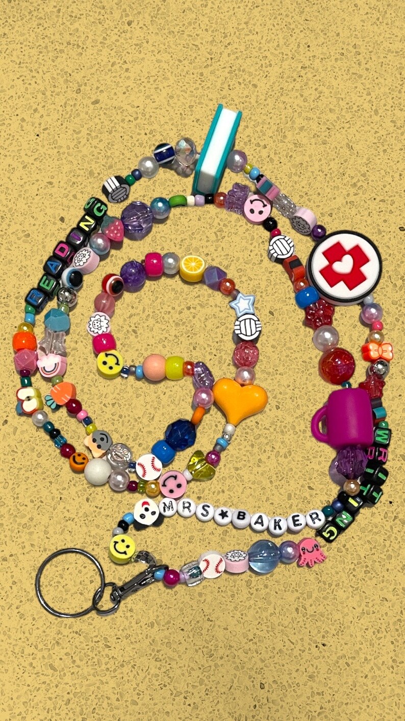 May include: A vibrant beaded lanyard with diverse charms, such as smiley faces, baseballs, and a miniature book. The lanyard spells out "READING" and "MRS BAKER" with letter beads. It includes a keyring and an assortment of beads in various shapes and colours.