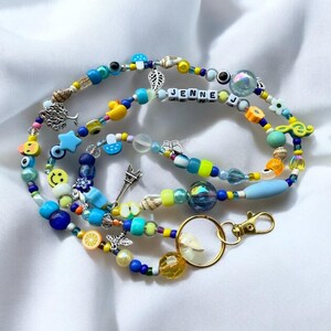 May include: A colourful beaded lanyard with a variety of beads, charms, and the name "JENNE" spelled out in white letter beads. The lanyard features a gold-coloured clasp and a mix of blue, yellow, and clear beads, along with charms like a tree, Eiffel Tower, and smiley faces.