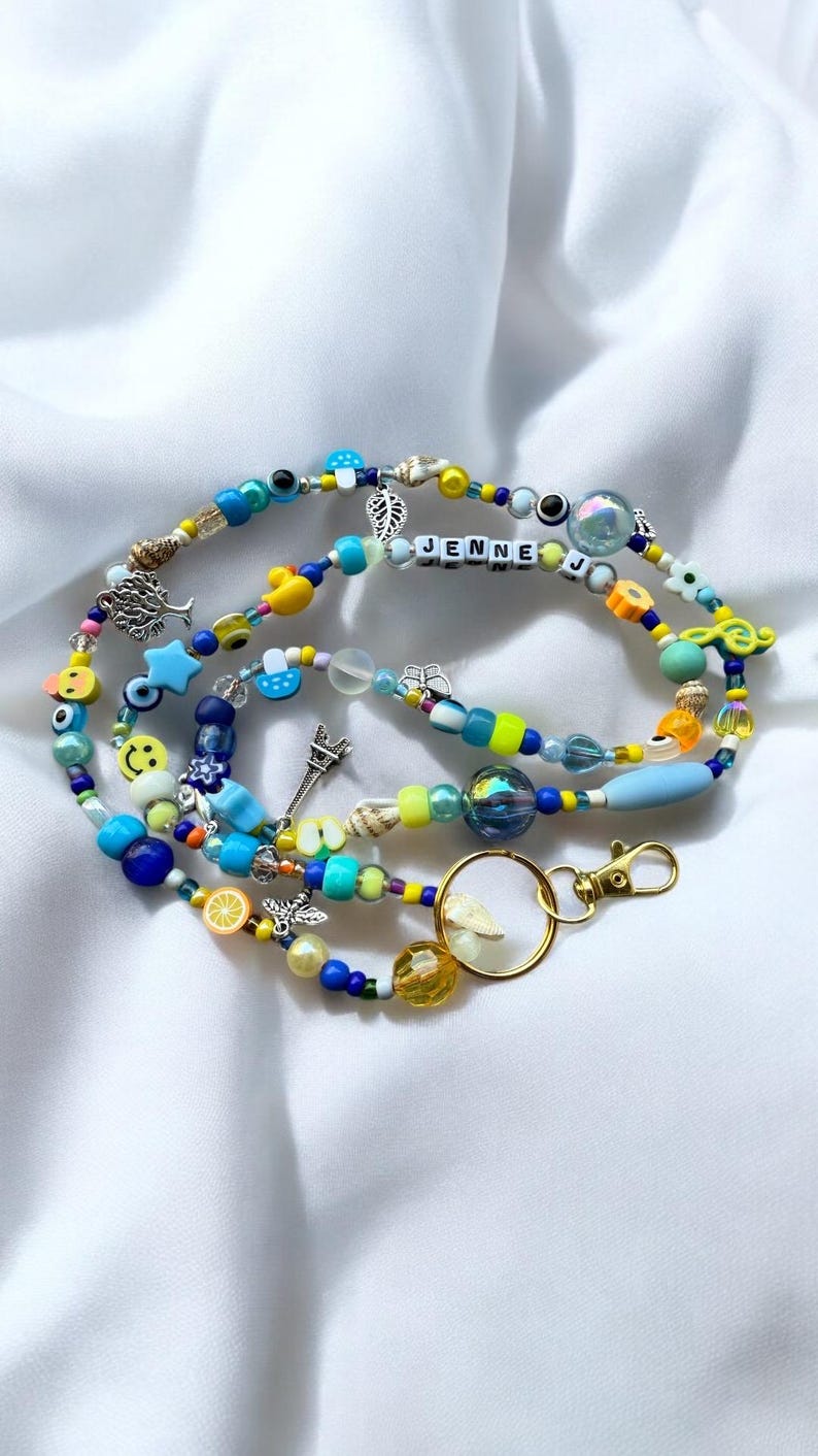 May include: A colourful beaded lanyard with a variety of beads, charms, and the name "JENNE" spelled out in white letter beads. The lanyard features a gold-coloured clasp and a mix of blue, yellow, and clear beads, along with charms like a tree, Eiffel Tower, and smiley faces.