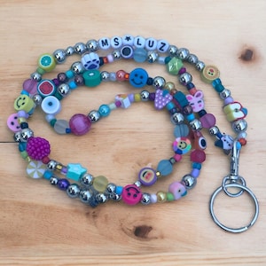 May include: A vibrant beaded lanyard with the name "MS LUZ" spelled out in letter beads. The lanyard features various beads, including smiley faces, fruit shapes, and star-shaped beads. It has a silver key ring attached.