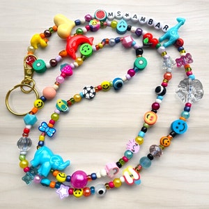 May include: A vibrant beaded lanyard featuring a variety of charms, such as smiley faces, fruit shapes, and a blue elephant. The lanyard includes the text "MS AMBAR" in white letter beads and a gold-coloured clasp.