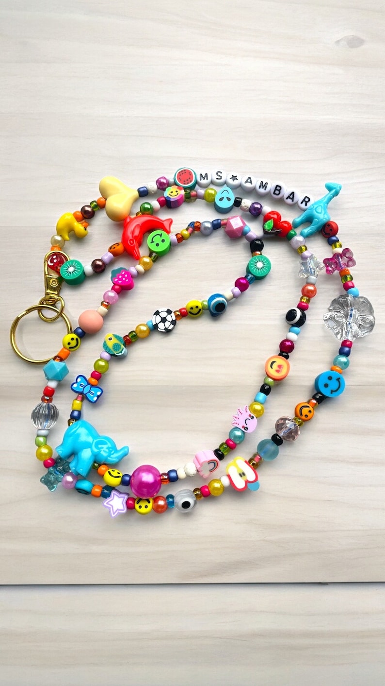 May include: A vibrant beaded lanyard featuring a variety of charms, such as smiley faces, fruit shapes, and a blue elephant. The lanyard includes the text "MS AMBAR" in white letter beads and a gold-coloured clasp.