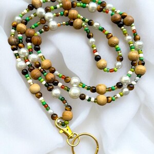 May include: A multi-strand beaded lanyard with wooden beads, faux pearls, and small colourful beads. It has a gold-coloured clasp and key ring. The beads are in shades of brown, white, green, and red.