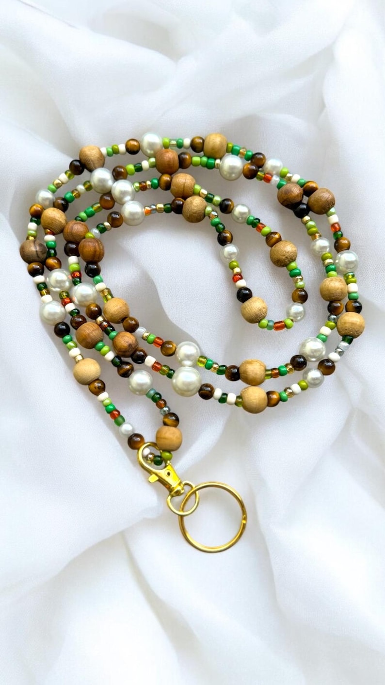 May include: A multi-strand beaded lanyard with wooden beads, faux pearls, and small colourful beads. It has a gold-coloured clasp and key ring. The beads are in shades of brown, white, green, and red.