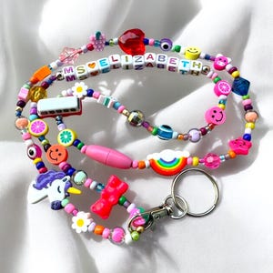 May include: A vibrant beaded lanyard with charms such as a rainbow, smiley faces, and a unicorn. The lanyard spells "ELIZABETH" with letter beads. It includes a lobster clasp and a key ring.