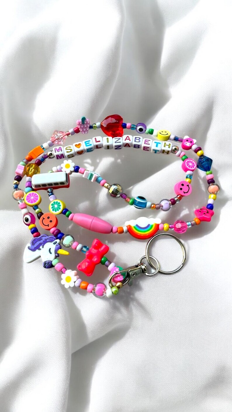 May include: A vibrant beaded lanyard with charms such as a rainbow, smiley faces, and a unicorn. The lanyard spells "ELIZABETH" with letter beads. It includes a lobster clasp and a key ring.