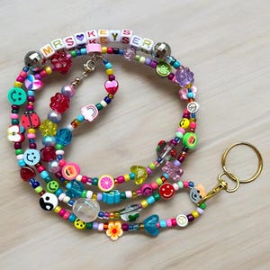 May include: A vibrant beaded lanyard with the text "MRS KEYS" and assorted charms. The lanyard includes smiley faces, hearts, flower and fruit-shaped beads. A gold-coloured key ring is attached.