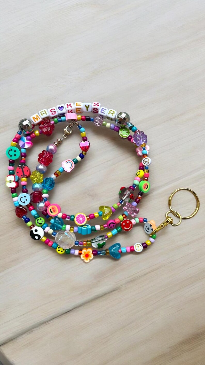 May include: A vibrant beaded lanyard with the text "MRS KEYS" and assorted charms. The lanyard includes smiley faces, hearts, flower and fruit-shaped beads. A gold-coloured key ring is attached.