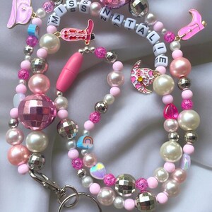 May include: A pink, white, and silver beaded lanyard with disco ball beads, pearls, and letter beads spelling "MISS NATALIE". The lanyard includes charms such as a pink boot, a pink ear, and a rainbow, all in a playful design.