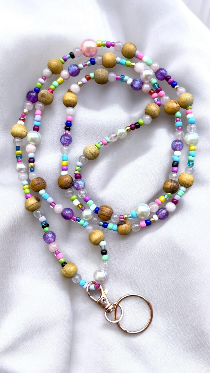 May include: A colourful beaded lanyard featuring wooden, iridescent, and pearl-like beads. The lanyard has a gold-coloured clasp and key ring. The beads are arranged in a layered design, creating a vibrant and eye-catching accessory.