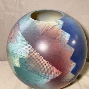 May include: A round ceramic vase with a blue, green and pink abstract design. The vase has a wide opening and a smooth, glossy finish.