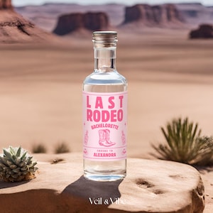 May include: A clear glass bottle with a pink label that says "Last Rodeo Bachelorette Cheers to Alexandra". The label has a pink illustration of cowboy boots. The bottle is sitting on a rock in a desert landscape.