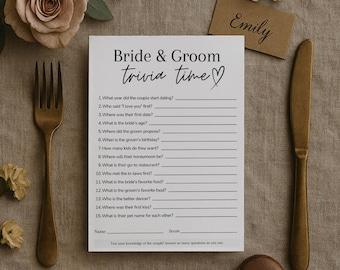 May include: A white Bride & Groom trivia game card with a heart, featuring questions about the couple. Includes a gold fork, knife, and a small card with the name "Emily". The card is surrounded by flowers.