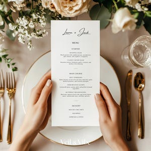 Könnte beinhalten: Eine Hochzeitsmenükarte, die von Händen gehalten wird und oben die Namen "Lauren + Jacob" trägt. Das Menü listet Vorspeisen, Hauptgerichte und Desserts auf. Goldenes Besteck und weiße Blumen umgeben die Karte und schaffen eine elegante, formelle Umgebung.