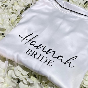 May include: White satin pajama top with black trim and the name "Hannah" and the word "BRIDE" printed in black script. The pajama top is resting on a bed of white flower petals.