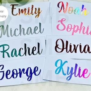May include: Eight rectangular white labels featuring custom names in various fonts and colours. The names displayed are Emily in gold glitter, Noah in holographic, Michael in silver, Sophia in pink, Rachel in green, Olivia in dark red, George in blue, and Kylie in a blue-to-pink gradient.